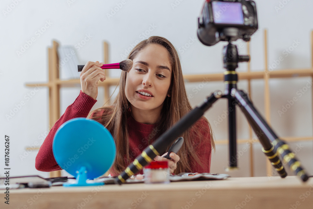Wonderful color. Attentive cheerful young blogger sitting in front of a mirror and putting on nice facial powder and recording it on a camera
