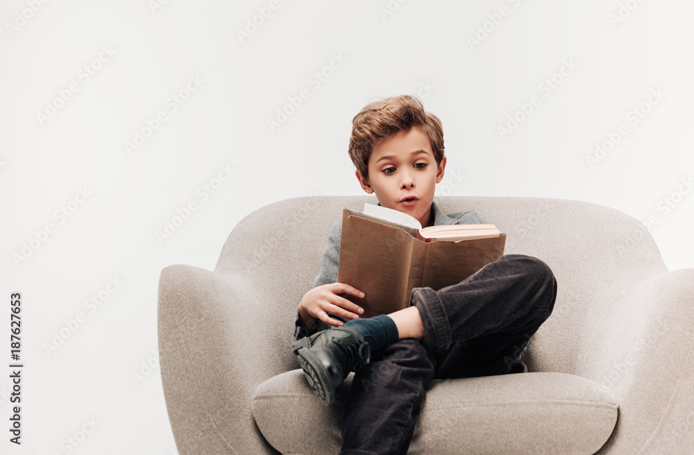 excited little schoolboy reading book in armchair isolated on grey excited little schoolboy reading book in armchair isolated on grey