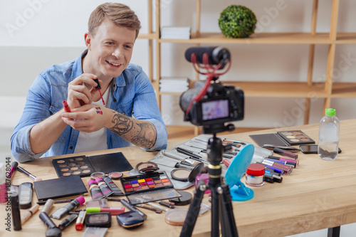 Blog recording. Young smart positive man feeling good while being surrounded by various cosmetics and recording a video blog about the usage of lipsticks