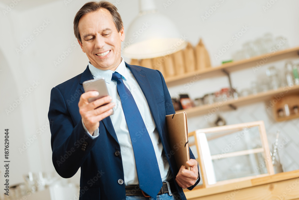 Good news. Senior occupied satisfied man standing in the room using the cellphone and holding the folder under left hand. Good news. Senior occupied satisfied man standing in the room using the cellphone and holding the folder under left hand.