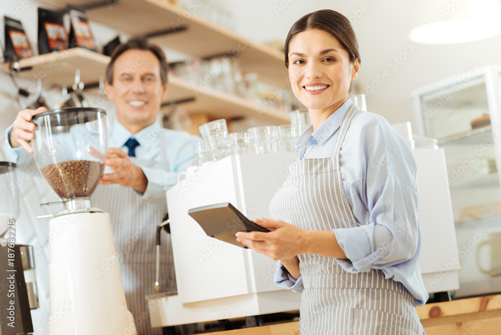 Qualified assistant. Beautiful pretty responsible worker standing near the coffee maker holding the calculator and smiling. Qualified assistant. Beautiful pretty responsible worker standing near the coffee maker holding the calculator and smiling.