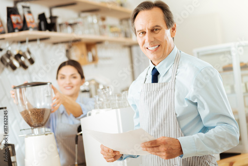 Simply work day. Handsome joyful satisfied man standing near the coffee machine smiling and holding the document.