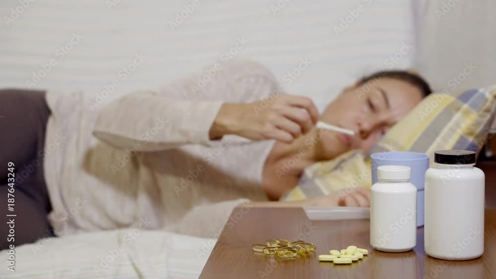 ill woman is lying on a sofa in a room, taking a thermometer out of mouth and taking antipyretic pill ill woman is lying on a sofa in a room, taking a thermometer out of mouth and taking antipyretic pill