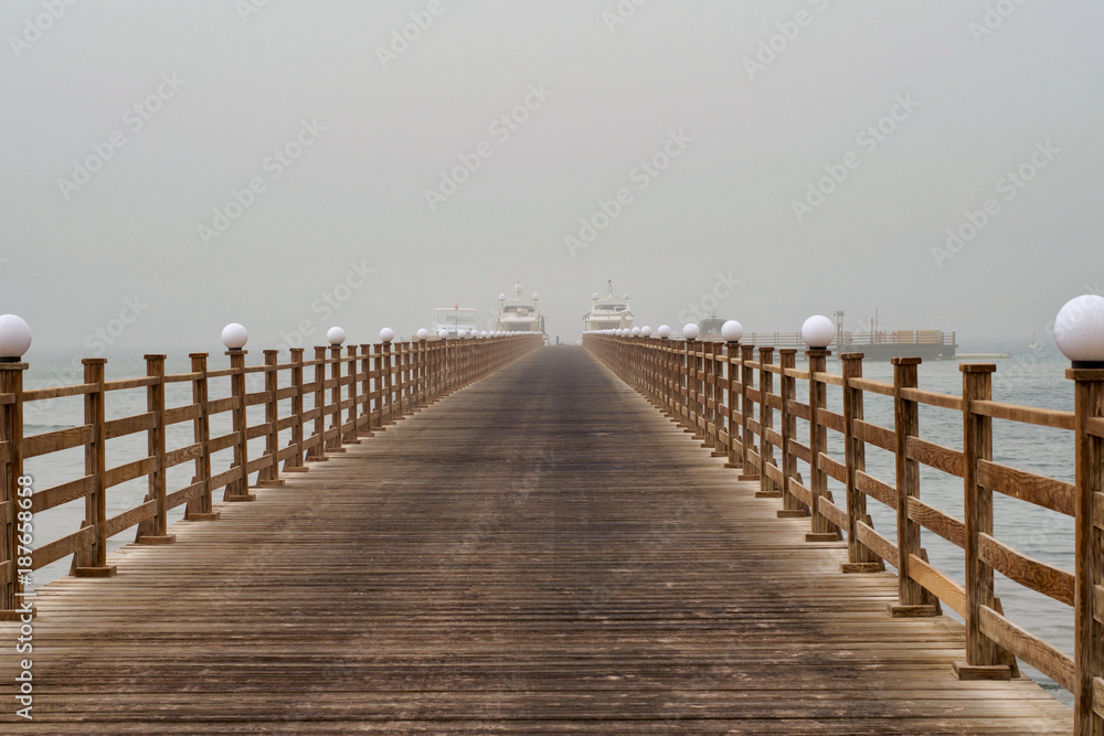 The wooden pier directs to the ships that stand at the pier in the fog. Natural blurring of the background.