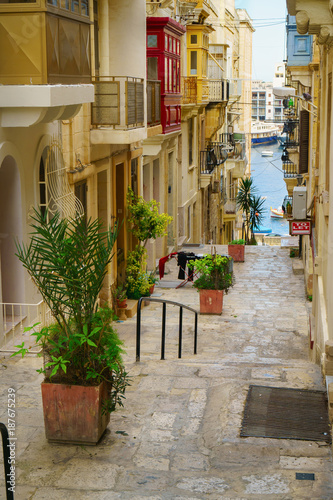 Fototapeta Naklejka Na Ścianę i Meble -  Medieval town narrow street with stairs in Valletta on the island of Malta. Colorful red, yellow, blue balconies and doors and green plants near traditional style stone buildings. Sea side view.