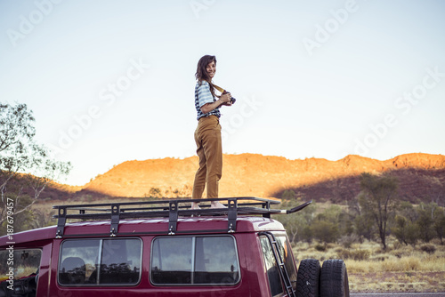 Smiling woman with camera standing on car roof against clear sky photo