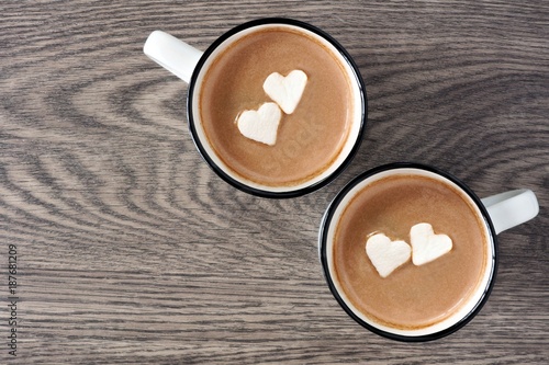 Two cups of hot chocolate with heart shaped marshmallows over a wooden background