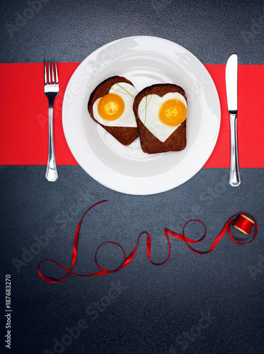Two toast with fried eggs in the shape of a heart on a white plate red ribbon bow.On dark marengo stone background.Inscription love of red satin ribbon.Creative Valentine day background.Love concept photo