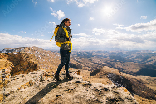 active young girl travels along the Caucasian ridge with a yellow backpack, stands on the edge of a cliff and enjoys the sun