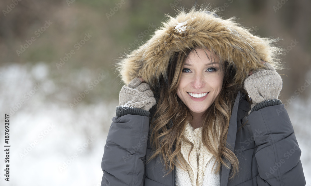 Obraz premium Beautiful brunette young woman in winter scene - snow covered park wearing winter clothes - layered sweaters and mittens holding park hood