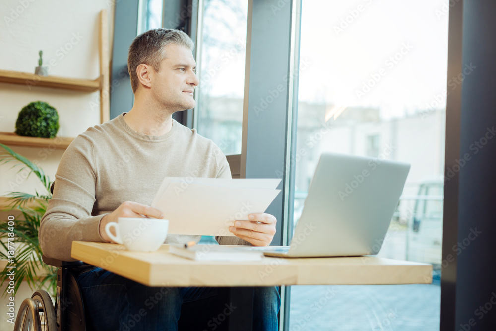 Thoughts. Handsome thoughtful handicapped man sitting in a wheelchair and holding some sheets of paper and looking out the window and working on his laptop in a cafe Thoughts. Handsome thoughtful handicapped man sitting in a wheelchair and holding some sheets of paper and looking out the window and working on his laptop in a cafe