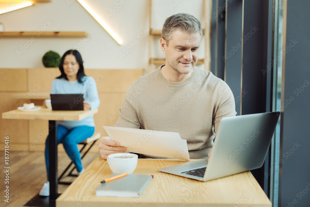 Working weekend. Attractive alert handicapped man sitting in a wheelchair and holding some sheets of paper and working on his laptop in a cafe and a woman sitting in the background Working weekend. Attractive alert handicapped man sitting in a wheelchair and holding some sheets of paper and working on his laptop in a cafe and a woman sitting in the background