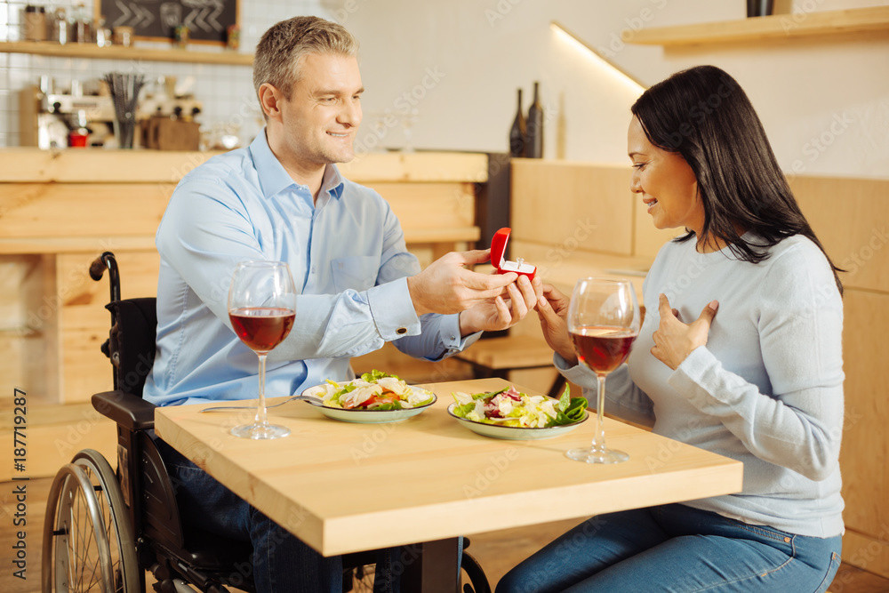 Be mine. Attractive inspired handicapped man smiling and proposing to his pretty beloved dark-haired woman and holding a ring while having romantic dinner