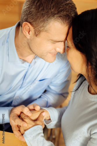 My precious. Inspired blond well-built man and a beautiful delighted dark-haired woman sitting with their eyes closed and holding hands and kissing