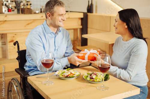 Present. Attractive content handicapped man smiling and giving a present to his pretty beloved alert woman while having romantic dinner