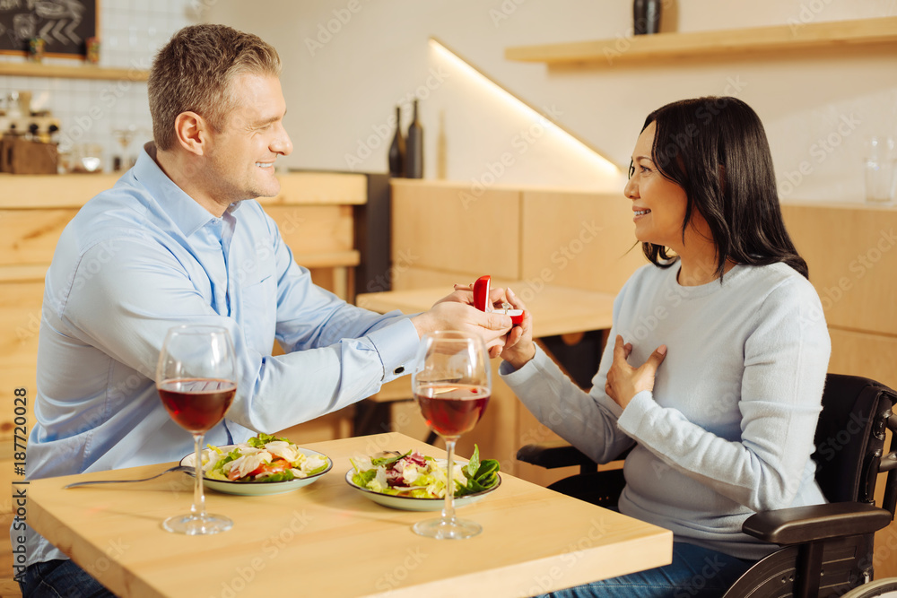 My bride. Good-looking gleeful well-built man smiling and proposing to his beautiful inspired handicapped woman while holding a ring and having dinner My bride. Good-looking gleeful well-built man smiling and proposing to his beautiful inspired handicapped woman while holding a ring and having dinner