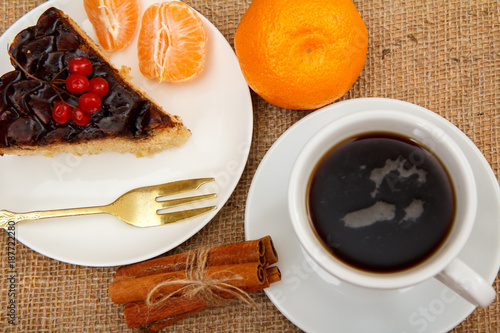 Slice of chocolate cake decorated with bunch of viburnum, fork, cup of coffee, oranges and cinnamon