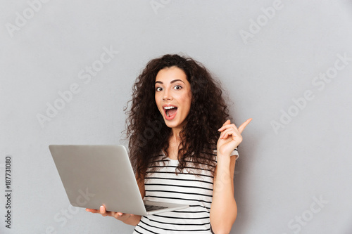 Studio portrait of woman with curly hair being excited to find useful information in internet via silver computer gesturing eureka with index finger photo