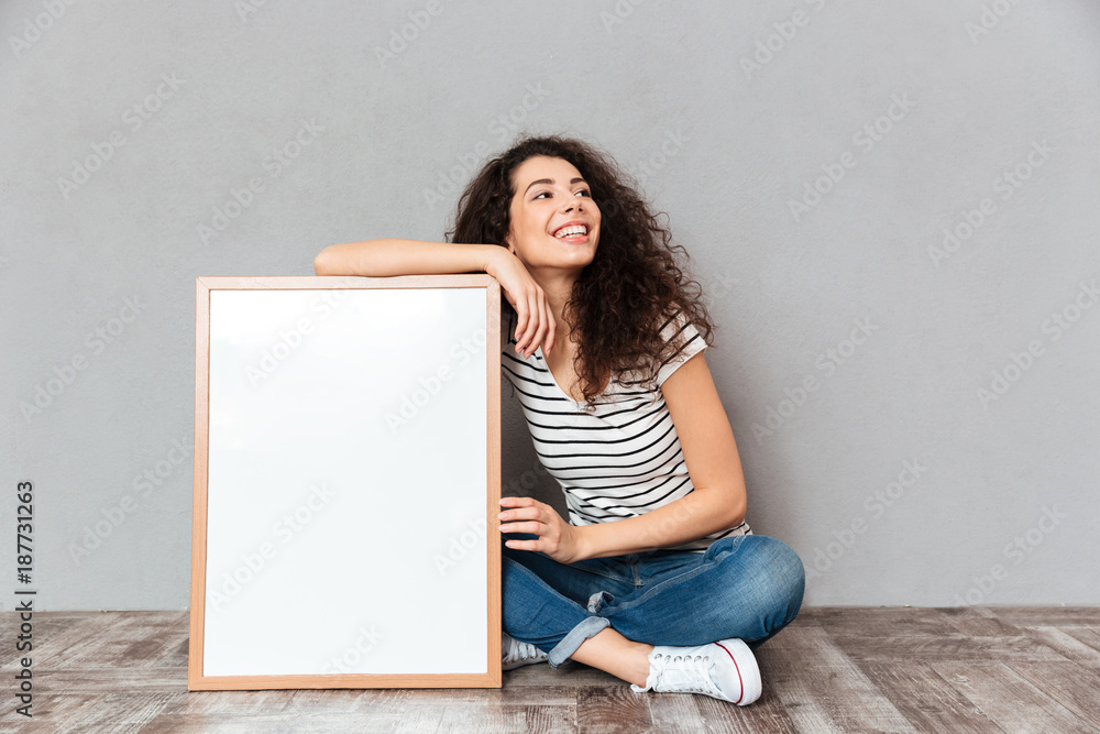 Naklejka premium Caucasian woman with beautiful hair posing with legs crossed demonstrating big great painting or portrait isolated over grey wall copy space