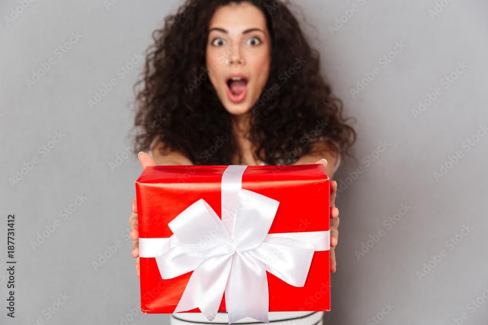 Brunette happy woman in defocus posing against grey wall holding red box gift-wrapped with white bow showing it on camera Brunette happy woman in defocus posing against grey wall holding red box gift-wrapped with white bow showing it on camera
