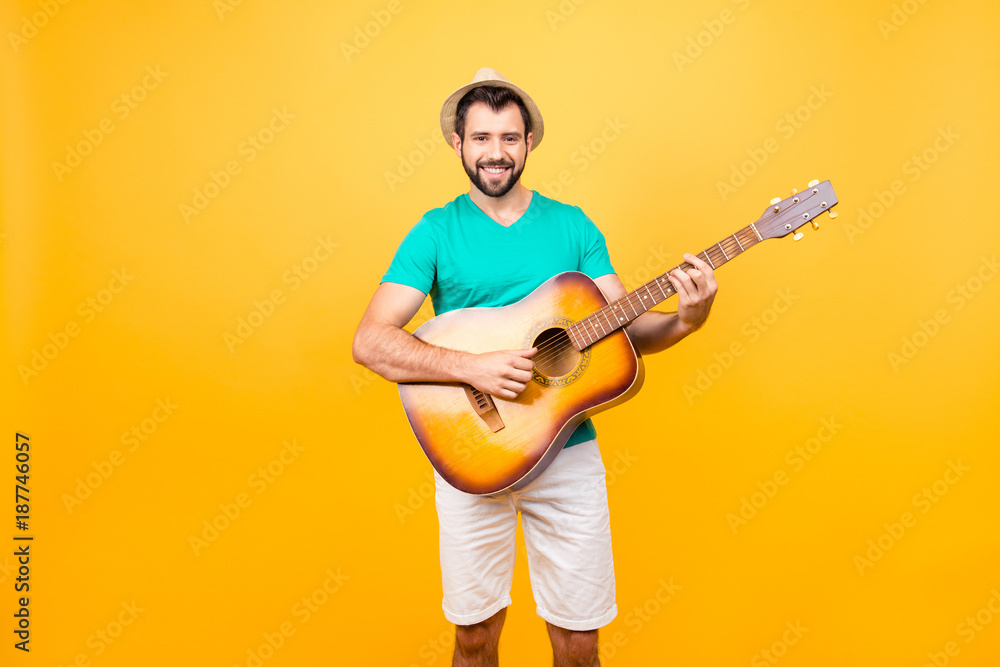 My guitar is my second soul! Portrait of smiling joyful funky glad positive guy with bristle, wearing tshirt and shorts, he is playing acoustic guitar, isolated on yellow background My guitar is my second soul! Portrait of smiling joyful funky glad positive guy with bristle, wearing tshirt and shorts, he is playing acoustic guitar, isolated on yellow background