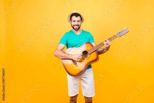 My guitar is my second soul! Portrait of smiling joyful funky glad positive guy with bristle, wearing tshirt and shorts, he is playing acoustic guitar, isolated on yellow background