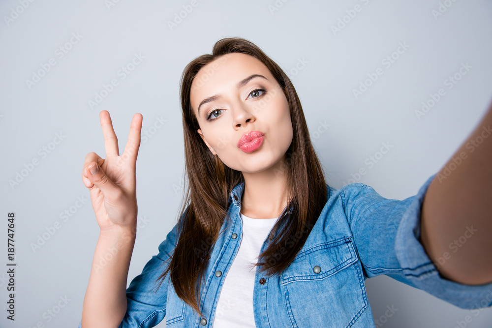 Self-portrait of confident brunette cute girl blowing kiss to the camera and gesturing peace symbol, standing over grey background Self-portrait of confident brunette cute girl blowing kiss to the camera and gesturing peace symbol, standing over grey background