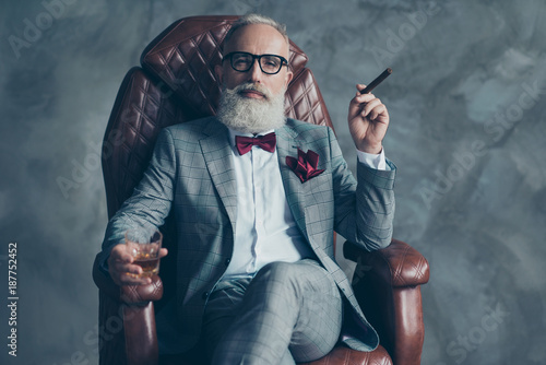 Cool man in glasses, hold cigarette,  glass with brandy, in formal wear, tux with red bowtie and pocket square, sit in leather chair over gray background, looking to the camera, shares, stock, money