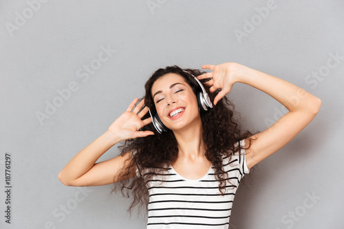 Picture of pleased young woman in striped t-shirt enjoying favourite songs listening melodies via headphones with closed eyes over grey background