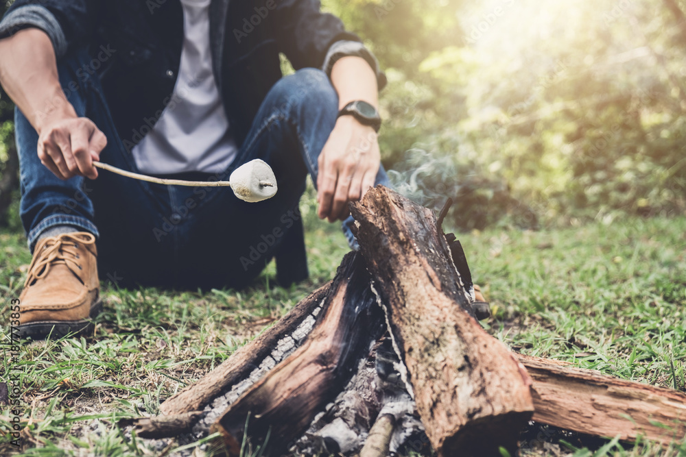 Obraz premium Young Man hiking cooking a marshmallow candies on the campfire in forest, hike and people concept - happy relaxation camping