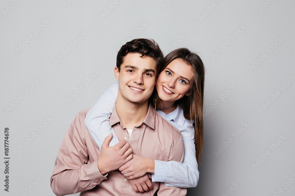 Portrait of a cheerful young couple hugging while standing and looking at camera over gray Portrait of a cheerful young couple hugging while standing and looking at camera over gray