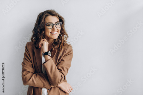 Emotional portrait of a beautiful and smiling girl in a glasses which stands near a white wall. Blank space for a text
