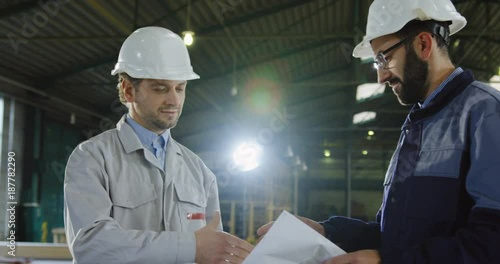 Factory worker and engineer in helmets with big paper drawings shaking hands and agreed on the project. Inside a big manufactory. Portrait shot. Close up photo