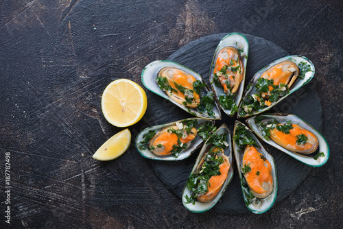 Boiled green mussels with parsley sauce and lemon served on a stone slate tray. Above view on a dark brown stone surface with copyspace