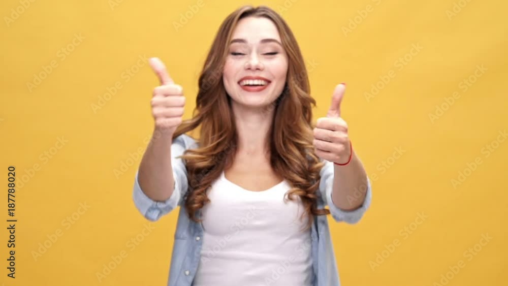 Happy brunette woman in denim shirt showing thumbs up and looking at the camera over yellow background Happy brunette woman in denim shirt showing thumbs up and looking at the camera over yellow background