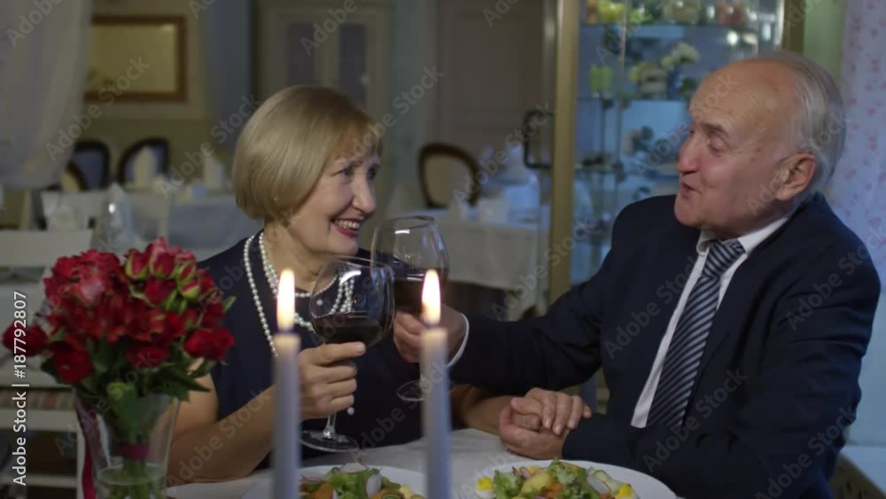 PAN of affectionate senior man in suit and happy elderly woman clinking glasses and drinking expensive wine on romantic date in restaurant PAN of affectionate senior man in suit and happy elderly woman clinking glasses and drinking expensive wine on romantic date in restaurant