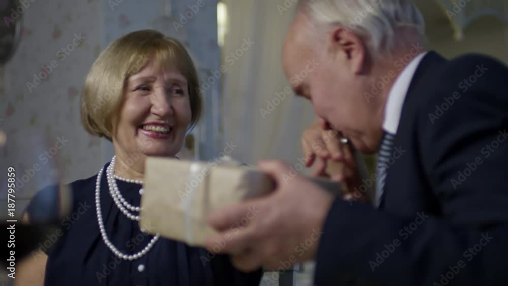 PAN of laughing senior woman giving present to affection elderly husband, then clinking glasses and drinking wine while celebrating anniversary in restaurant PAN of laughing senior woman giving present to affection elderly husband, then clinking glasses and drinking wine while celebrating anniversary in restaurant