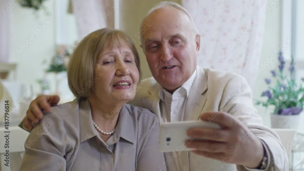 Medium shot of laughing senior woman and elderly man taking selfie and pulling funny faces while having date in cafe Medium shot of laughing senior woman and elderly man taking selfie and pulling funny faces while having date in cafe