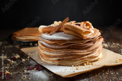 two stacks of pancakes, two folded pancakes, sprinkled with berries, cookies, sugar powder and caramel syrup on parchment on a dark wooden table