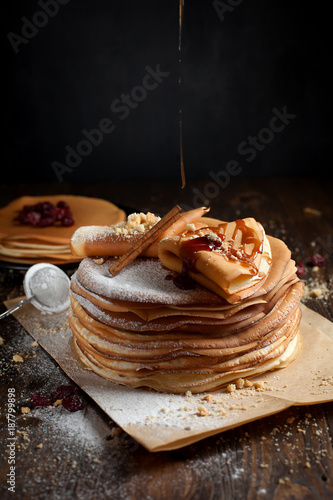 two stacks of pancakes, two folded pancakes, sprinkled with berries, cookies, sugar powder and caramel syrup on parchment on a dark wooden table