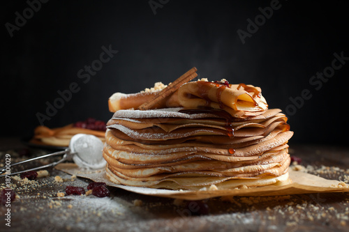two stacks of pancakes, two folded pancakes, sprinkled with berries, cookies, sugar powder and caramel syrup on parchment on a dark wooden table