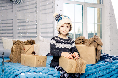 little girl the child sitting in pajamas and hat on the bed with garland of light bulbs with gifts boxes wrapped in a non-colored paper decorated with cones on blue knitted coverlet.Christmas concept photo