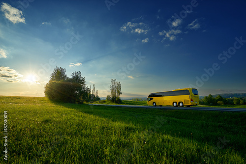 Obraz na płótnie Yellow bus traveling on the road between green meadows in a rural landscape at sunset w salonie