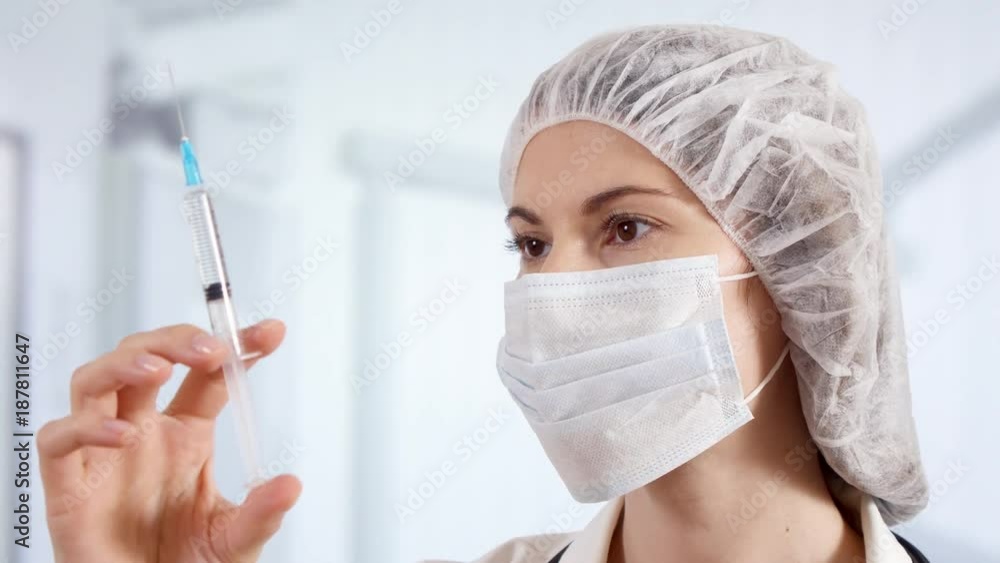 Professional female doctor in mask, cap and gloves in hospital room with disposable one-off syringe with transparent drug. Woman physician at work. Laboratory employee making scientific research Professional female doctor in mask, cap and gloves in hospital room with disposable one-off syringe with transparent drug. Woman physician at work. Laboratory employee making scientific research