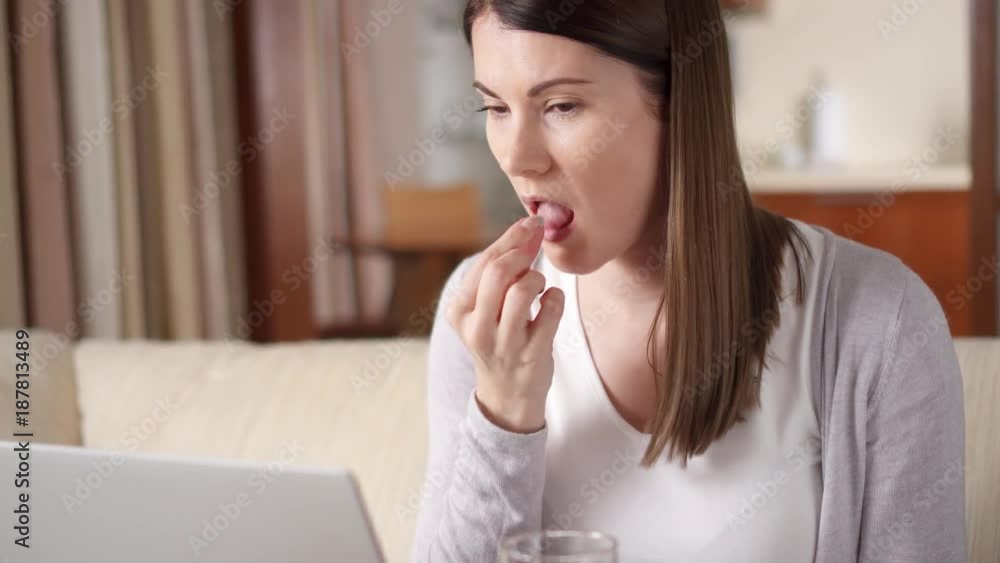 Unwell young businesswoman with severe headache taking her medication at home. Professional female felling sick ill migraine having painkiller pills drugs with glass of water Unwell young businesswoman with severe headache taking her medication at home. Professional female felling sick ill migraine having painkiller pills drugs with glass of water