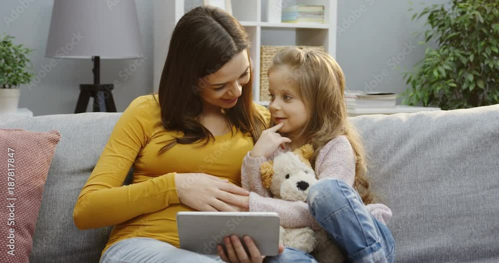 Portrait shot of the charming pregnant woman sitting close with her cute little daughter, holding a tablet device and showing to her how baby kicking inside in the cozy living room. Indoors Portrait shot of the charming pregnant woman sitting close with her cute little daughter, holding a tablet device and showing to her how baby kicking inside in the cozy living room. Indoors