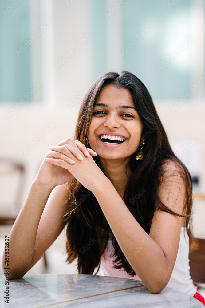 Portrait of young and attractive Indian woman in a white top sitting in a  wood chair and smiling radiantly Stock Photo
