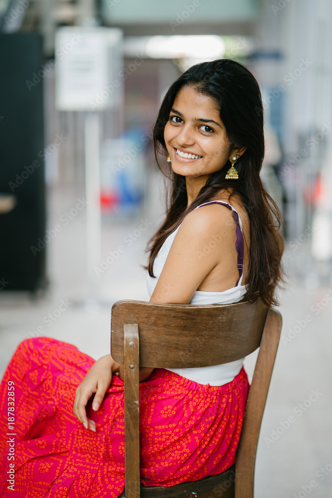 Portrait of young and attractive Indian woman in a white top sitting in a  wood chair and smiling radiantly Stock Photo