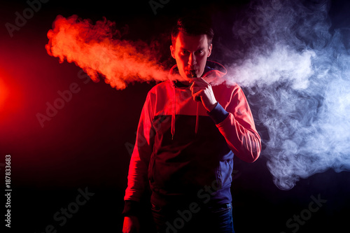 Young stylish male smoker in a sweatshirt is smoking a vape, and around a big cloud of smoke from vape on a dark isolated background