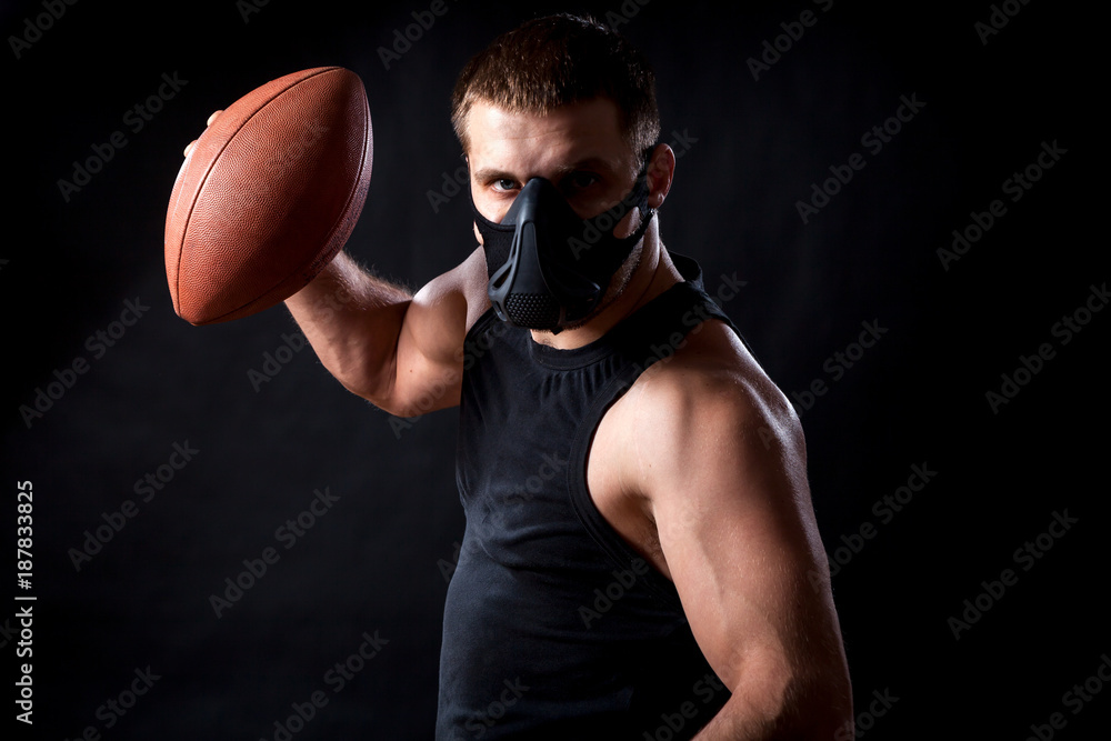 Portrait of young athletic man wearing sport shirt, training mask and holding rugby ball on black isolated background Portrait of young athletic man wearing sport shirt, training mask and holding rugby ball on black isolated background